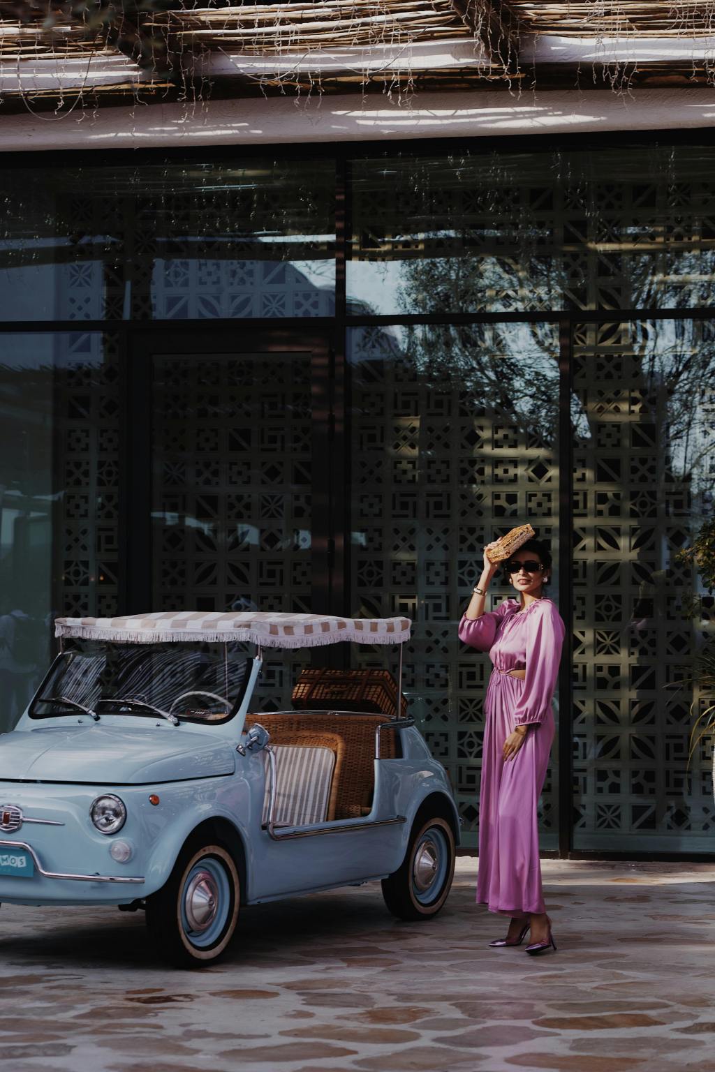 A stylish woman wearing a flowing pink maxi dress poses beside a pastel-blue vintage convertible with wicker accents, showcasing elegant retro fashion for the modern AI-inspired homemaker.