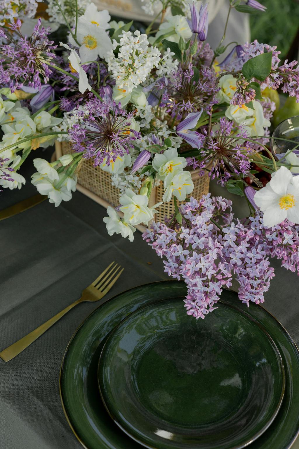 Spring garden party tablescape featuring a woven basket filled with lilacs, white anemones, and purple alliums, set on a dark tablecloth with gold flatware and green ceramic plates, creating a fresh and elegant outdoor dining atmosphere.